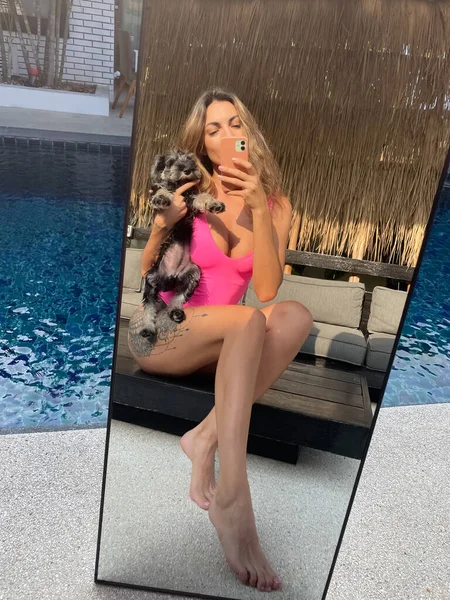 在户外 穿着亮晶晶粉色泳衣的体态丰满的 晒得发亮的女人在户外的社交媒体 游泳池边与小狗垂直拍摄自己的照片 — 图库照片
