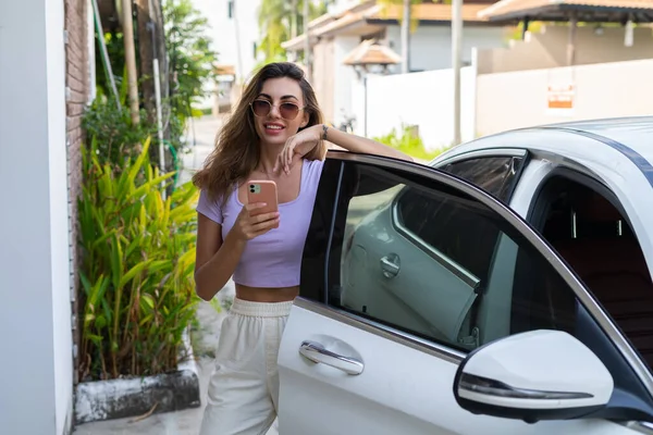 カジュアルな服装で成功した笑顔フィット日焼けした魅力的な女性は 屋外の高級近代的な車の近くに立っている間 彼女のスマートフォンを使用しています — ストック写真