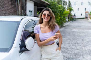 Başarılı gülümseyen, bronzlaşmış, gündelik giyimli çekici bir kadın lüks modern arabanın yanında dururken akıllı telefonunu kullanıyor..