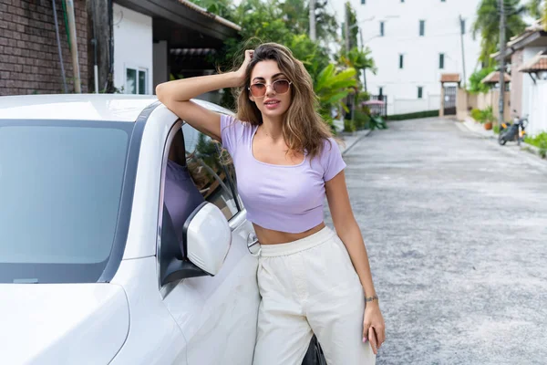 カジュアルな服装で成功した笑顔フィット日焼けした魅力的な女性は 屋外の高級近代的な車の近くに立っている間 彼女のスマートフォンを使用しています — ストック写真