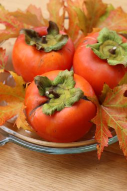 Tahta bir masada sonbahar yapraklı cam bir kasede olgun kaki ya da Persimmon meyveleri. Diospyros kaki meyveleri