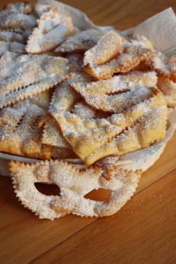 Maske şeklinde İtalyan Karnavalı pastası. Geleneksel karnaval tatlı kıtır börek kızartılmış ve üzerine pudra şekeri serpiştirilmiş. Chiacchere veya crostoli, bugie, sanci beyaz bir tabakta ahşap masada 