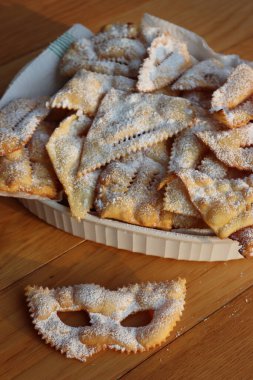 Maske şeklinde İtalyan Karnavalı pastası. Geleneksel karnaval tatlı kıtır börek kızartılmış ve üzerine pudra şekeri serpiştirilmiş. Chiacchere veya crostoli, bugie, sanci beyaz bir tabakta ahşap masada 