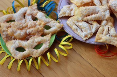 Italian sweet crisp pastry deep-fried and sprinkled with sugar in shape of Carnival mask on colorful plates. Chiacchere or crostoli, bugie, cenci with confetti and paper serpentine  clipart