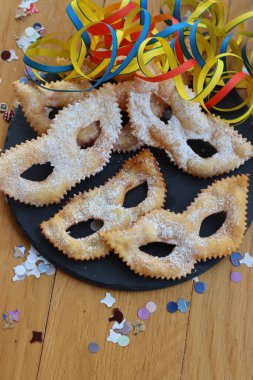 Traditional sweet crisp pastry deep-fried in shape of Carnival mask on a black slate stone plate. Chiacchere or crostoli, bugie, cenci with confetti and paper serpentine 