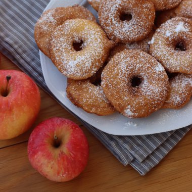 Homemade deep fried apple rings with batter sprinkled with powdered sugar ona white plate on wooden table with fresh apples