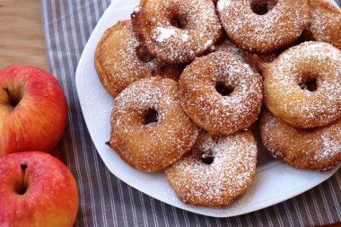 Homemade deep fried apple rings with batter sprinkled with powdered sugar ona white plate on wooden table with fresh apples