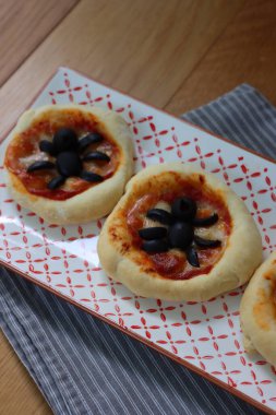  Ahşap masadaki seramik tepside siyah zeytinli Cadılar Bayramı minik örümcek pizzaları.