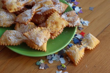 Masada yeşil bir tabakta fiyonk şeklinde kızartılmış İtalyan tatlısı. Chiacchere veya crostoli, bugie, renkli kağıt konfetiyle cenci 