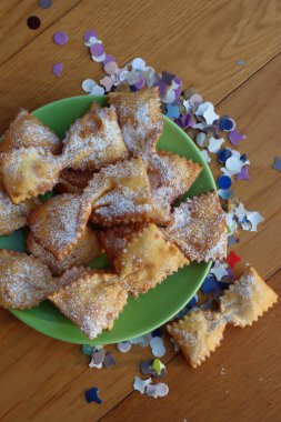 Masada yeşil bir tabakta fiyonk şeklinde kızartılmış İtalyan tatlısı. Chiacchere veya crostoli, bugie, renkli kağıt konfetiyle cenci 