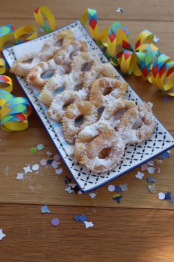 Italian sweet crisp pastry deep-fried and sprinkled with sugar in shape of Carnival mask on a tray. Chiacchere or crostoli, bugie, cenci with confetti and paper serpentine clipart