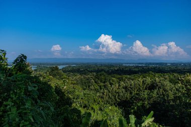 Bereketli yeşil ormanlarla kaplı tepelerin üzerinde süzülen bulutlu gökyüzünün manzarası. Bu sakin manzara, doğal güzelliği, barışçıl atmosferi ve el değmemiş vahşi doğanın canlı yeşilliğini vurgular..