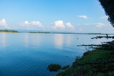 Serene Kaptai Gölü, berrak mavi bir gökyüzünün altında uzak tepeler ve sakin suda parıldayan yumuşak bulut yansımaları. Huzurlu bir manzara, seyahat fotoğrafçılığı, doğa severler ve turizm keşfi için mükemmel..
