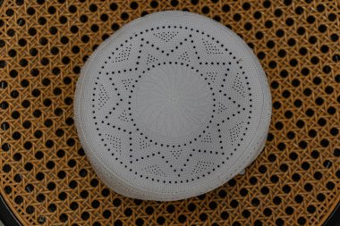 Zarif, beyaz işlemeli dua şapkası dokunmuş kamış koltuk yüzeyine düzgünce yerleştirilir. İslami moda, dini kıyafetler, ruhani aksesuarlar, Müslüman kültürü, dua ritüelleri ve törensel başlık kavramları için ideal stok imajı..