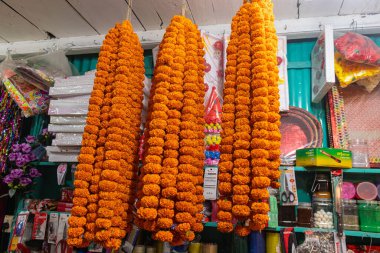 Parlak turuncu kadife çiçekli çelenkler canlı bir pazar ortamında asılı duruyor, taze geleneksel çiçek süslemeleri, festival renkleri, kültürel sembolizm, doğal güzellik, kutlama, Hint pazarı sahnesi, yakın çekim dokusu ve aromatik çiçekler sergileniyor..