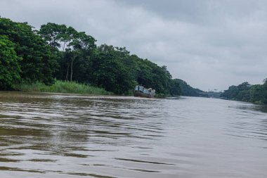 A faded painted cargo boat glides past lush green river islands beneath heavy grey monsoon clouds, capturing the calm, humid atmosphere of river life during the rainy season.