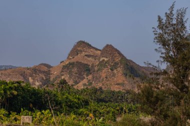 Görkemli bir kahverengi kayalık tepe, berrak mavi bir kıyı gökyüzünün altında gür bir tropikal palmiye ormanının üzerinde yükselir, doğal güzellikleri ve sakin tropikal manzaraları gözler önüne serer..
