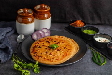 Aloo paratha, baharatlı patates püresiyle doldurulmuş lezzetli bir Hint ekmeği, keskin turşu (achar) ve ferahlatıcı baharatla sıcak servis edilir, mükemmel bir lezzet ve sıcaklık dengesi yaratır..