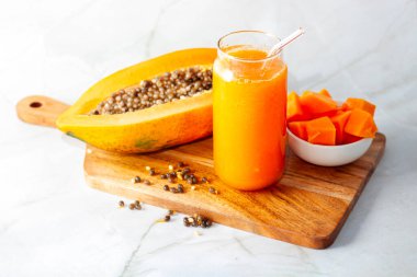 A halved papaya, vibrant smoothie in a glass jar with a straw, fresh cubes in a bowl, and scattered seeds on a wooden board clipart