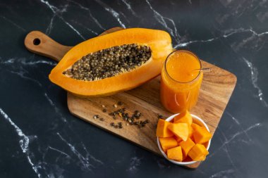 A halved papaya, vibrant smoothie in a glass jar with a straw, fresh cubes in a bowl, and scattered seeds on a wooden board clipart