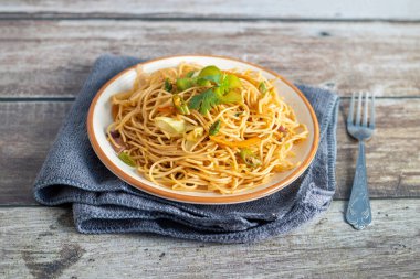 Canlı bir tabak taze kişniş ve sebzeli kızarmış şehriye, kıvrımlı bir ahşap çatalın yanında katlanmış gri bir peçetenin üzerinde duruyor..