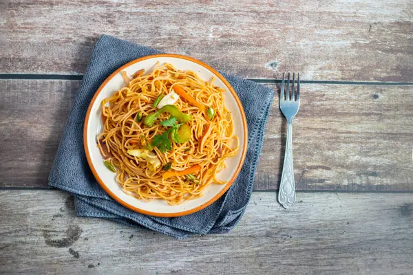 Canlı bir tabak taze kişniş ve sebzeli kızarmış şehriye, kıvrımlı bir ahşap çatalın yanında katlanmış gri bir peçetenin üzerinde duruyor..