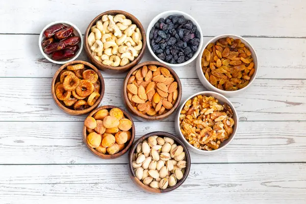 Kurumuş meyve ve fındık çeşitleri beyaz ahşap bir yüzeye yerleştirilmiş, hurma, kaju, badem, kuru üzüm, incir, kayısı ve ceviz de dahil olmak üzere..