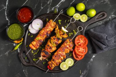 Yanında doğranmış domates, soğan, misket limonu ve sosla servis edilen demir bir tavada sebzeli ızgara tavuk şişleri..