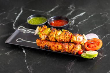 Yanında doğranmış domates, soğan, misket limonu ve sosla servis edilen demir bir tavada sebzeli ızgara tavuk şişleri..