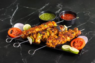 Yanında doğranmış domates, soğan, misket limonu ve sosla servis edilen demir bir tavada sebzeli ızgara tavuk şişleri..