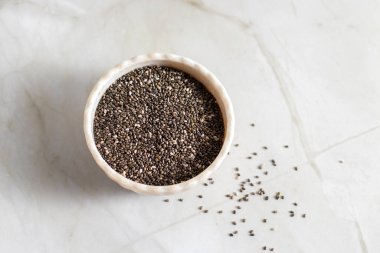 Siyah chia tohumlarıyla dolu beyaz bir seramik kase hafif bir mermer yüzeyde duruyor, bazı tohumlar kaselerin etrafına saçılmış durumda..