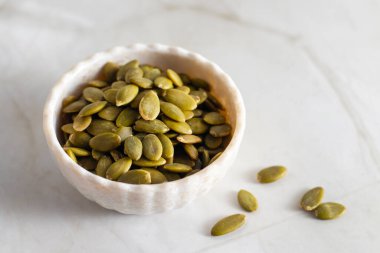 Yeşil balkabağı tohumlarıyla dolu beyaz bir seramik kase hafif bir mermer yüzeyde duruyor ve birkaç tohum kasenin etrafına saçılmış durumda..