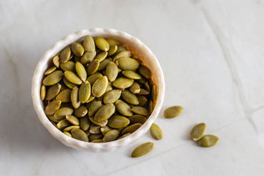 Yeşil balkabağı tohumlarıyla dolu beyaz bir seramik kase hafif bir mermer yüzeyde duruyor ve birkaç tohum kasenin etrafına saçılmış durumda..