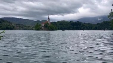 Slovenya 'daki Bled Gölü' nün ortasında güzel dağlar ve küçük bir ada. Bulutlu yağmurlu hava, su yansıması, yüzen tekne. Yüksek kalite 4k görüntü