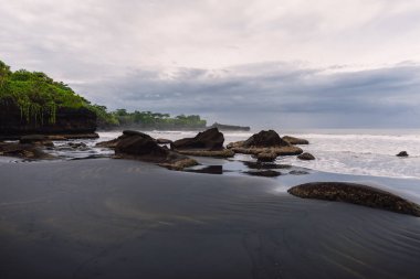 Balian, Bali 'de okyanuslu ve dalgalı volkanik kumlu plaj. Sahilde kayalar ve bulutlu gökyüzü