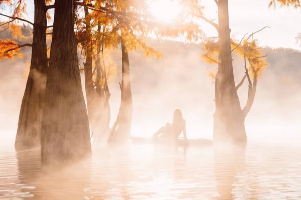 Woman relaxing on paddle board at the lake with morning fog, sun and autumnal Taxodium distichum trees
