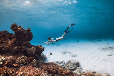 Bikinili serbest dalgıç kız, mavi okyanusta tropikal balıklarla mercan resifi yakınlarında yüzgeçleriyle süzülüyor.