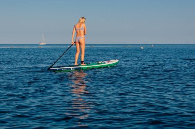 30 Temmuz 2022. Budva, Karadağ. Sıska sarışın kadın, sakin denizde sabah ışığıyla ayakta kürek çekiyor. Okyanusta Red Paddle SUP sörf tahtasında yüzen kadınlar.