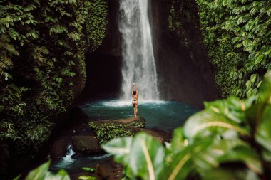 Scenic waterfall and woman in bikini near waterfall. Traveler girl posing on waterfall
