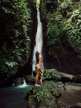 Slim woman posing on rock near waterfall. Traveler girl posing on scenic waterfall