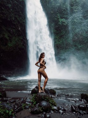 Beautiful slim woman in bikini posing near waterfall in tropical Bali. Traveler girl posing on river with powerfull waterfall