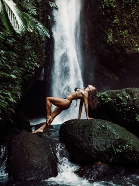 Gorgeous slim woman in bikini posing near waterfall. Traveler girl posing on waterfall