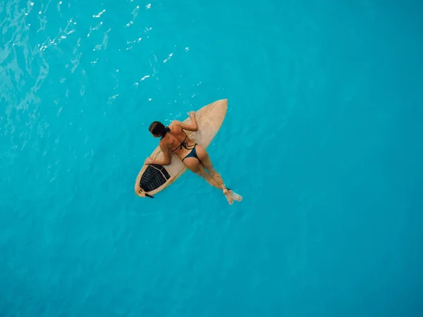 Surf girl in bikini swimming with surfboard in turquoise ocean. Aerial view of surfer woman