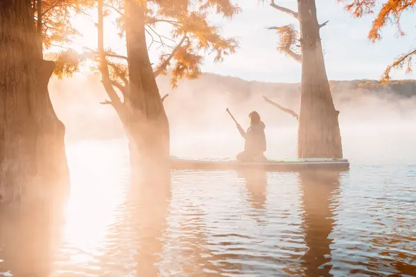 Woman relaxing on staand up paddle board at quiet lake with morning fog and fall Taxodium distichum trees