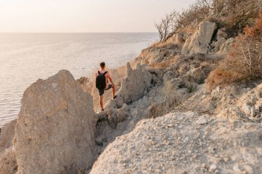 Tişört giyen ve sırt çantalı kısa şortlu bir yürüyüşçü, sıcak güneş ışığı olan kayaların arasında kıyıda kalıyor..