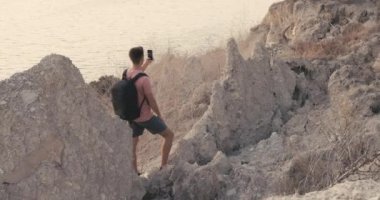 Okyanus kıyısında, sıcak güneş ışığıyla kayaların arasında kalan yürüyüşçü. Gezgin adam dışarıda akıllı telefonla video çekiyor..