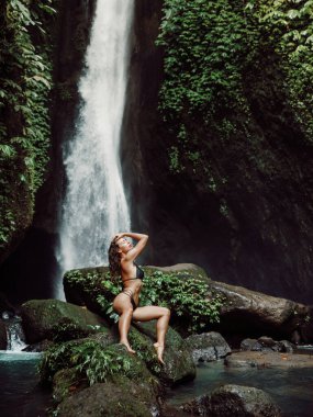 Bikinili zayıf kadın şelalenin yanında eğleniyor. Şelalede poz veren genç bir kız.
