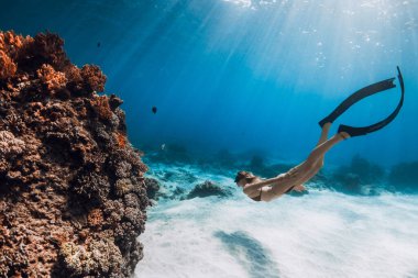 Bikinili kadın, tropikal mavi denizde mercanların yanında yüzgeçleriyle suyun altında yüzer.