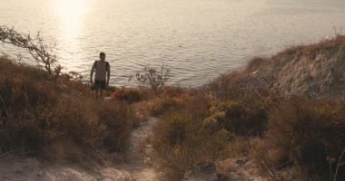 Tişörtlü ve şortlu bir yürüyüşçü. Sırt çantalı. Kıyı şeridinde sıcak gün batımı ışığı altında yürüyor. Yüksek kalite 4k görüntü