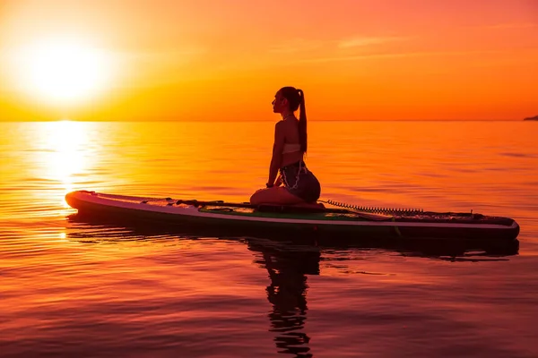 Slim young woman sitting on stand up paddle board at sea with bright sunset or sunrise. Woman relax on sup board in quiet sea.